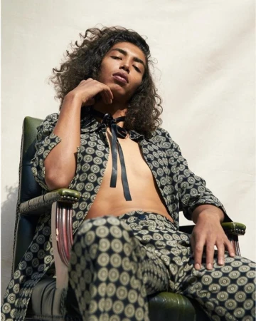 Male fashion model with curly hair poses in patterned pants and jacket, seated in a green chair in an indoor setting with natural light.