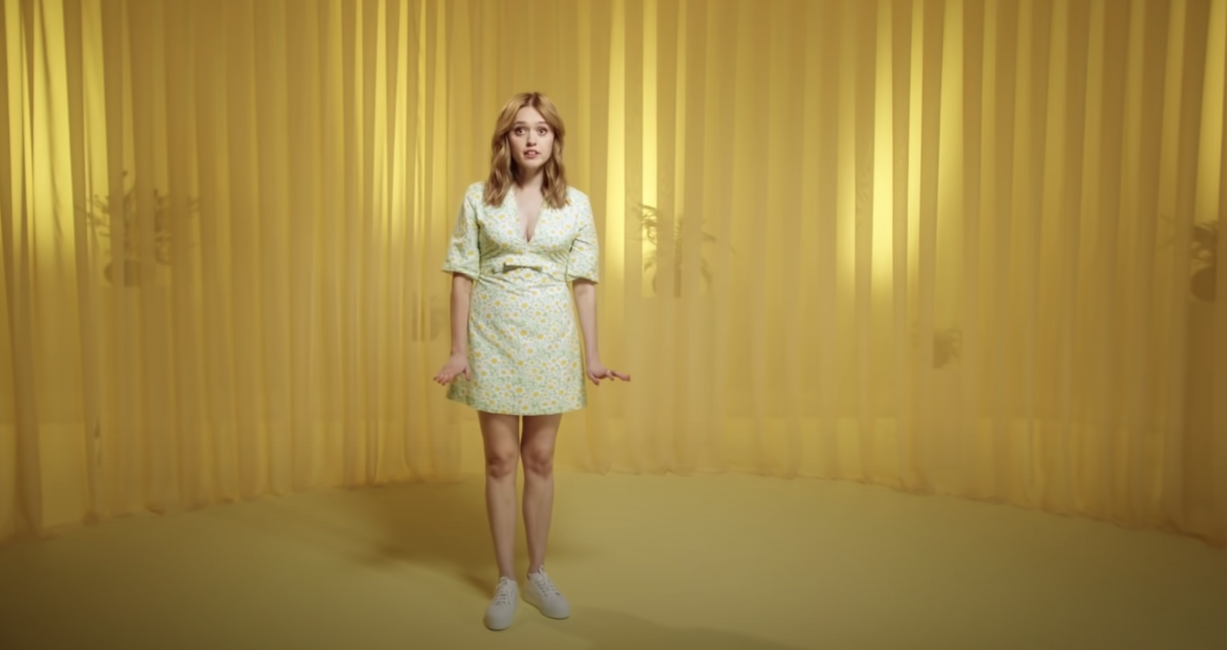 Full-length shot of adult female in floral dress and white sneakers against yellow draped backdrop in brightly lit photo studio.