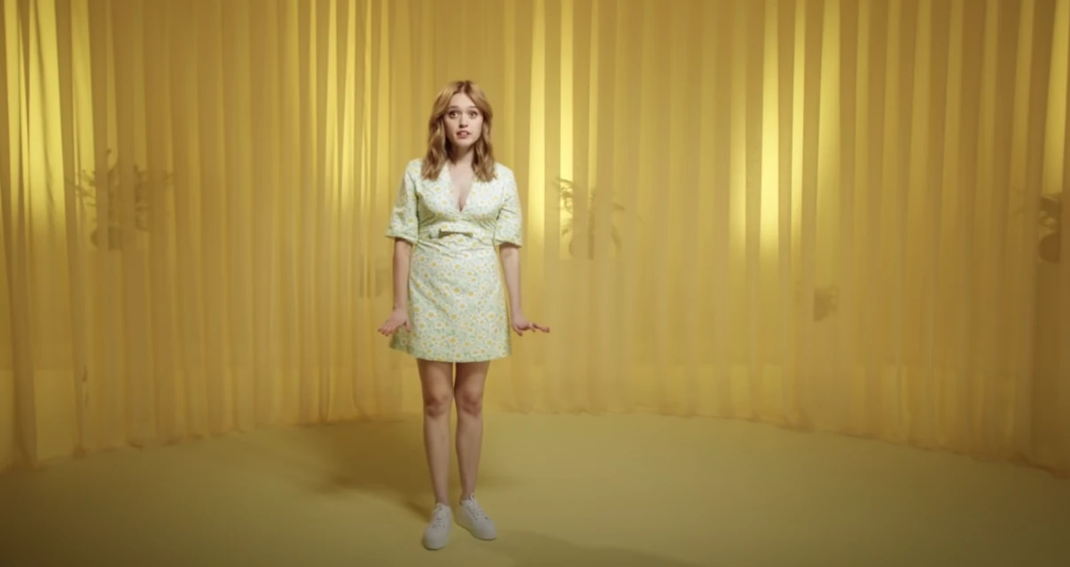 Full-length shot of adult female in floral dress and white sneakers against yellow draped backdrop in brightly lit photo studio.