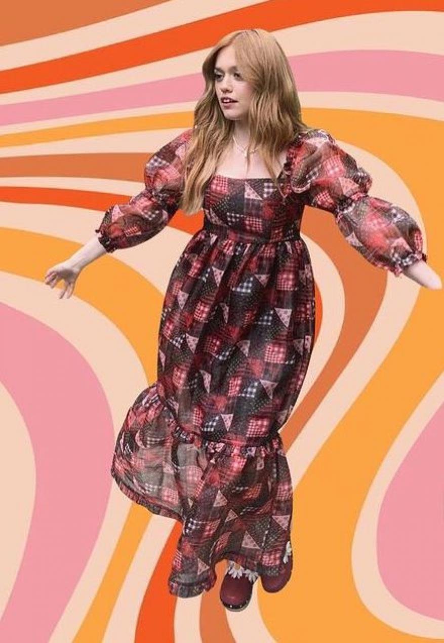 A woman wearing a vintage-inspired patterned dress with puff sleeves poses against a wavy, retro-style background in a full shot.
