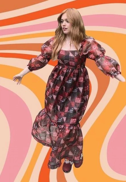 A woman wearing a vintage-inspired patterned dress with puff sleeves poses against a wavy, retro-style background in a full shot.