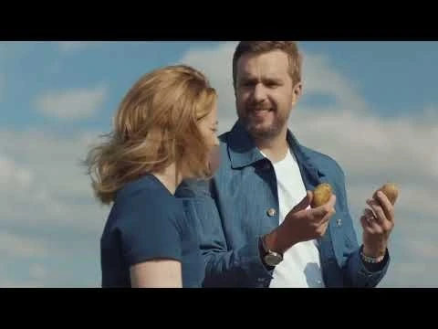 A smiling man and woman stand outdoors under a blue sky, holding potatoes. The man wears a blue jacket over a white t-shirt.