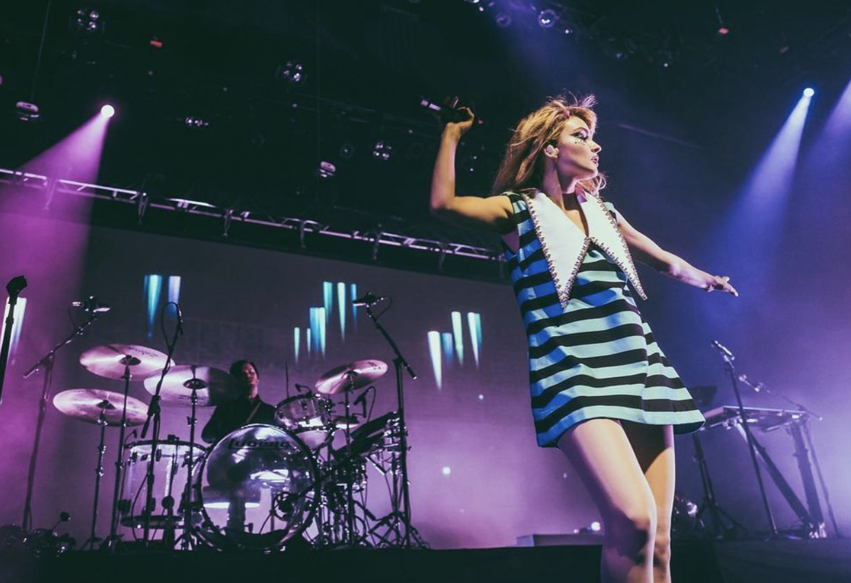 Female singer performing live on stage in a striped dress, backed by a drummer and keyboard. Purple stage lighting creates a vibrant atmosphere.