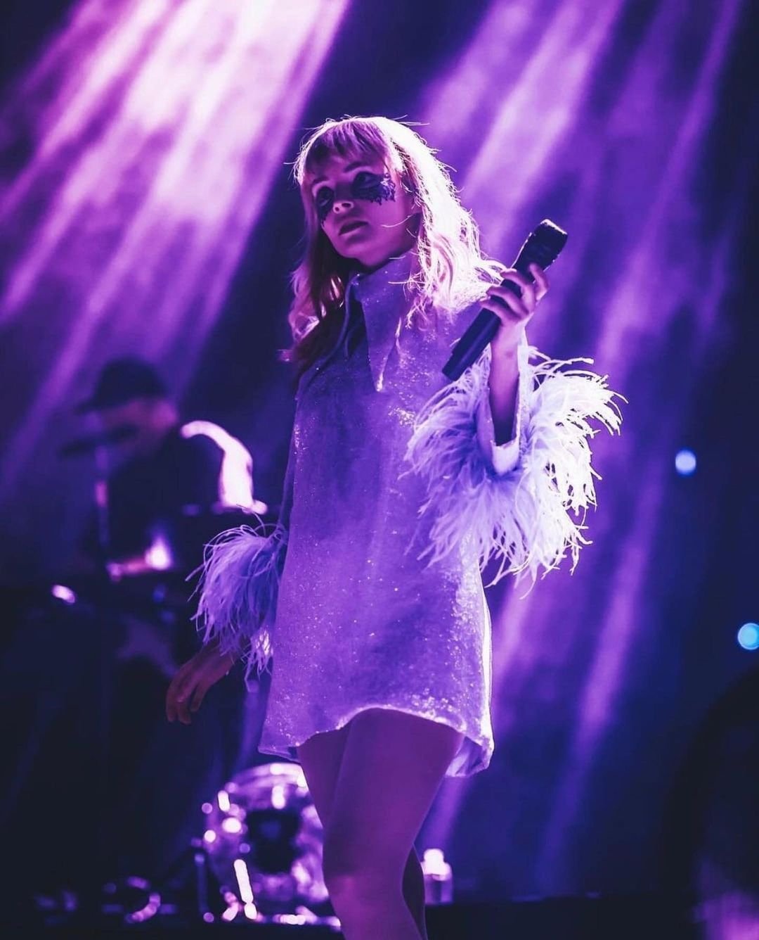 A singer performing on stage with a microphone in hand, wearing a shiny short dress with feathered sleeves, with stage lighting.