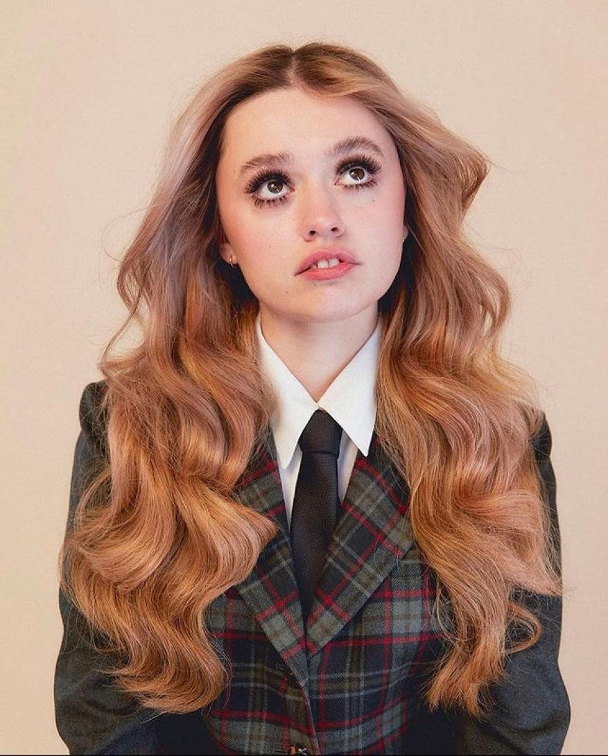 Young woman with long wavy blonde hair wearing a plaid blazer, white shirt, and black tie against a neutral background