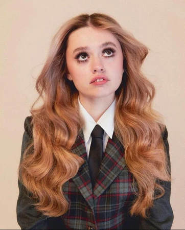 Young woman with long wavy blonde hair wearing a plaid blazer, white shirt, and black tie against a neutral background
