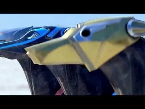 Close-up of two futuristic, multi-colored vehicles with black tires against a blurred sandy background under a bright, cloudless sky.