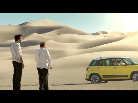Two men stand in a desert landscape with sand dunes and a yellow SUV visible in the background, suggesting a travel or automotive theme.