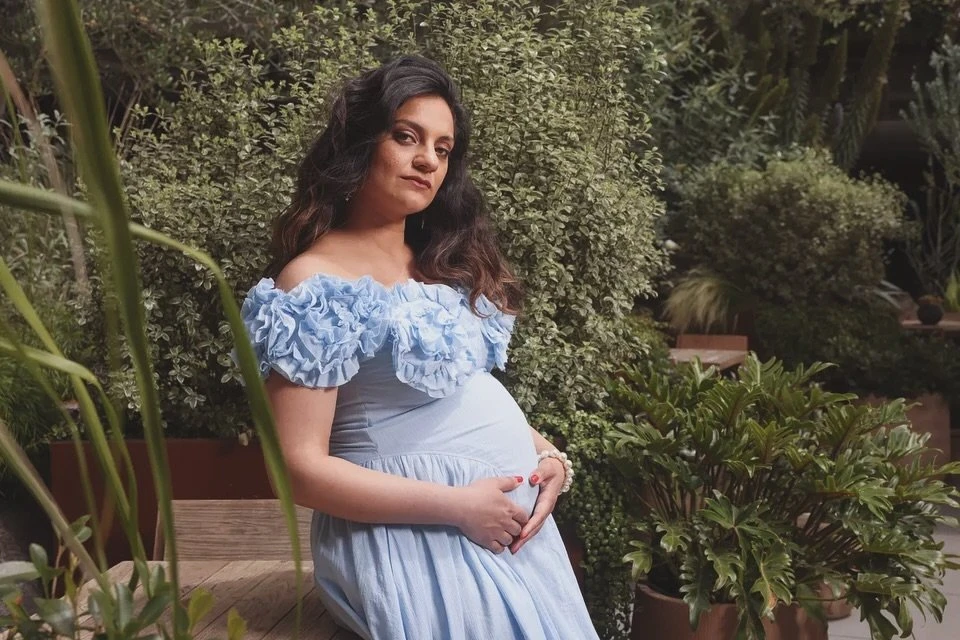 Pregnant woman in light blue dress sits outdoors surrounded by lush greenery, cradling her baby bump in a serene maternity portrait.