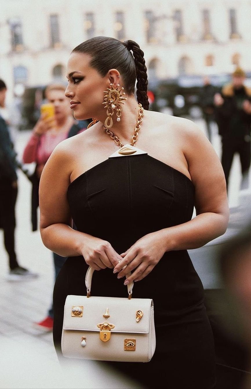 Woman in black dress with cream-colored purse and gold jewelry at an outdoor event, side-view.