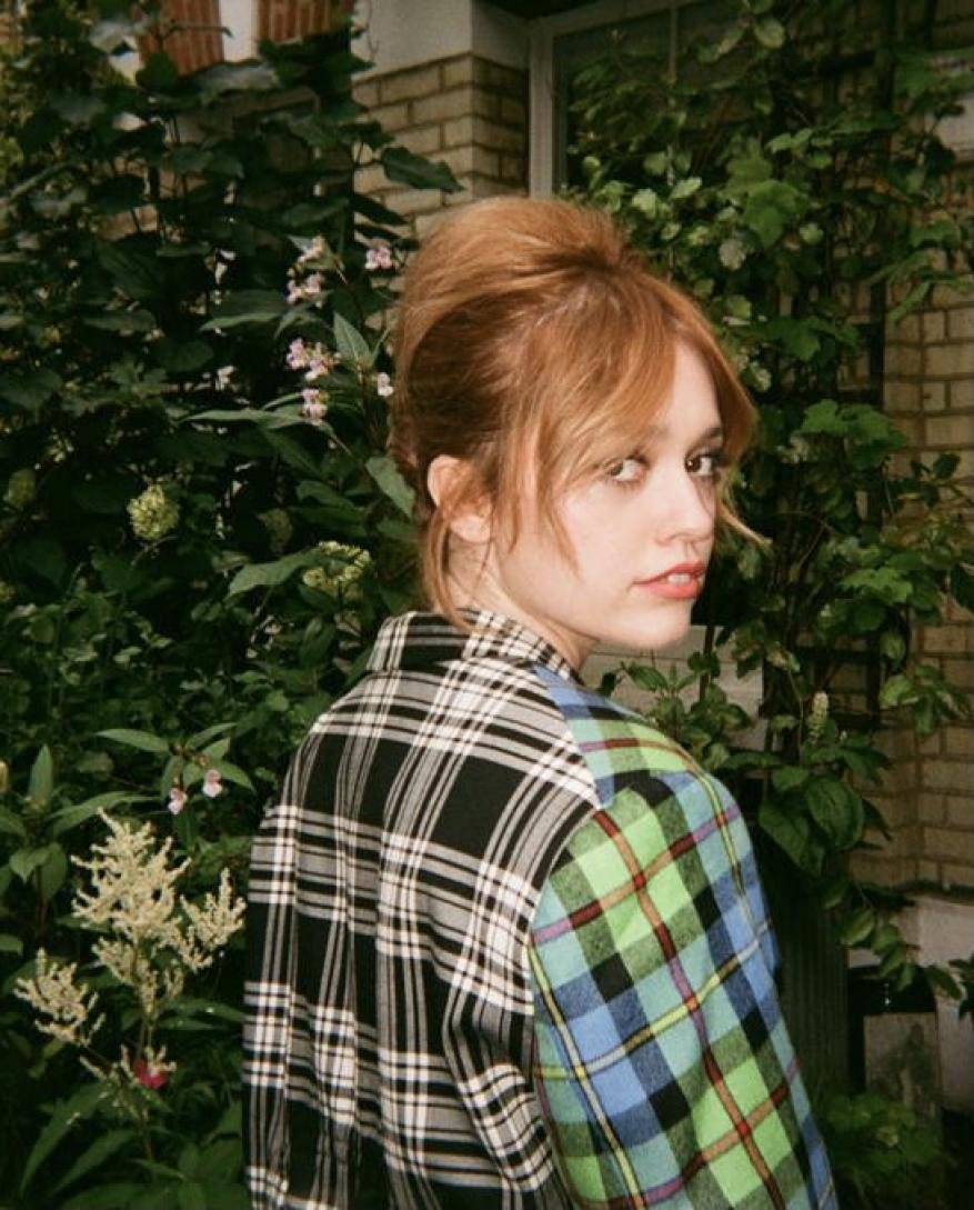 Portrait of a young adult woman with auburn hair and a plaid shirt, set against a background of leafy greenery and a brick building.