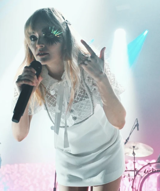 A woman with blond hair, wearing a white dress, sings into a microphone on a stage with spotlights and a band in the background.