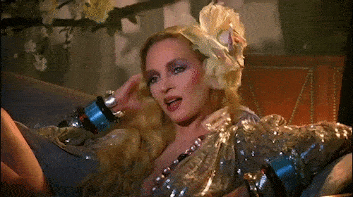 Woman with blond hair reclining on a couch wearing a gold sequin outfit, flower hair piece, bracelets, and dark eye makeup.
