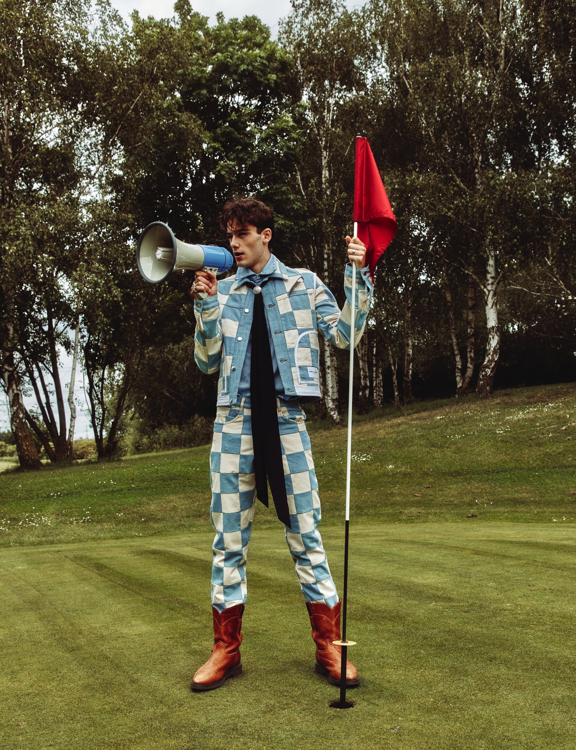 A young adult male stands on a manicured golf course, wearing a patchwork suit and holding a megaphone and golf flag with a red flag.