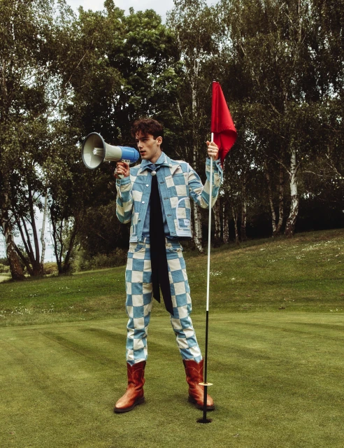 A young adult male stands on a manicured golf course, wearing a patchwork suit and holding a megaphone and golf flag with a red flag.