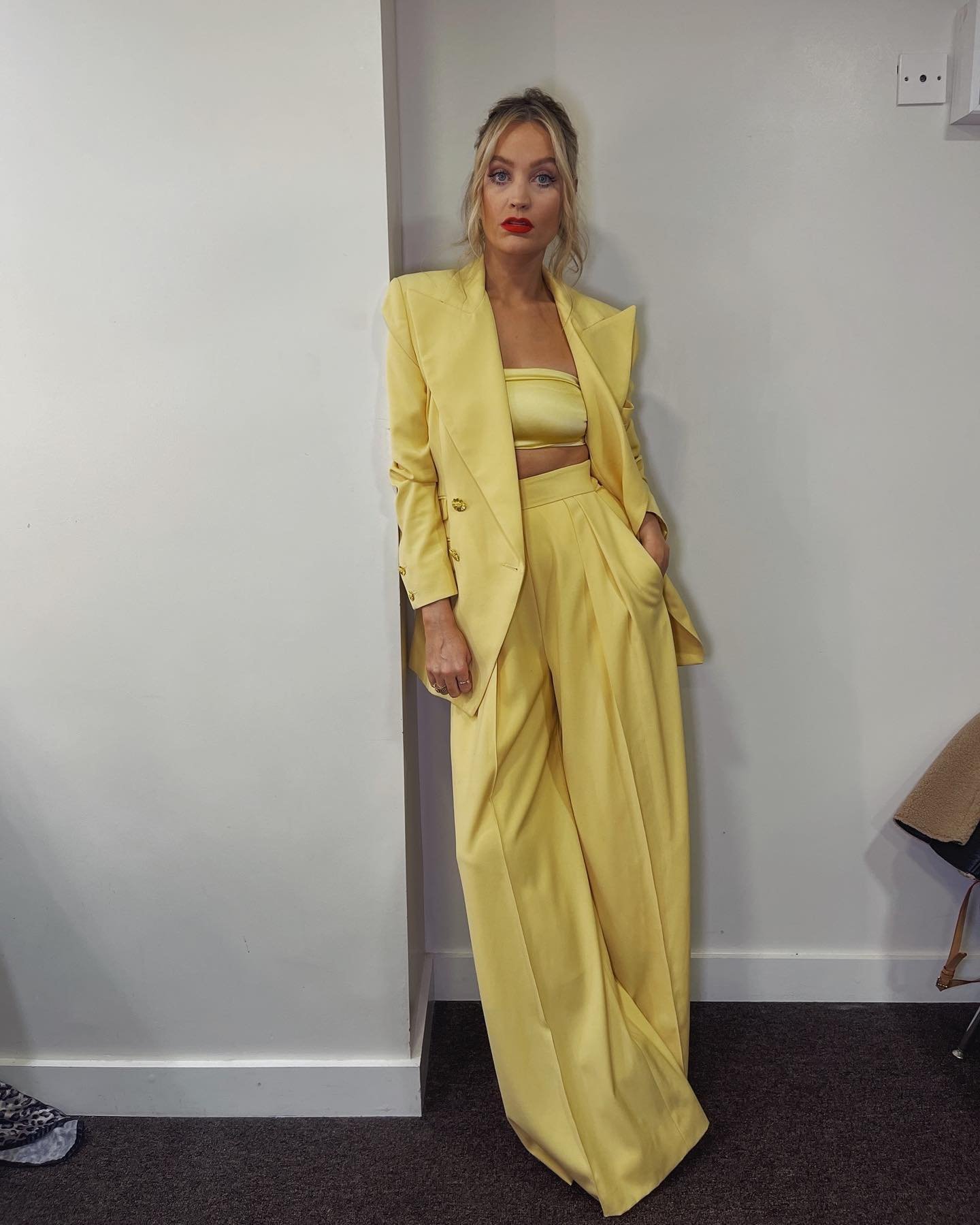 A blonde adult female model poses indoors against a white wall wearing a light-yellow-colored blazer, tube top, and wide-leg pants.