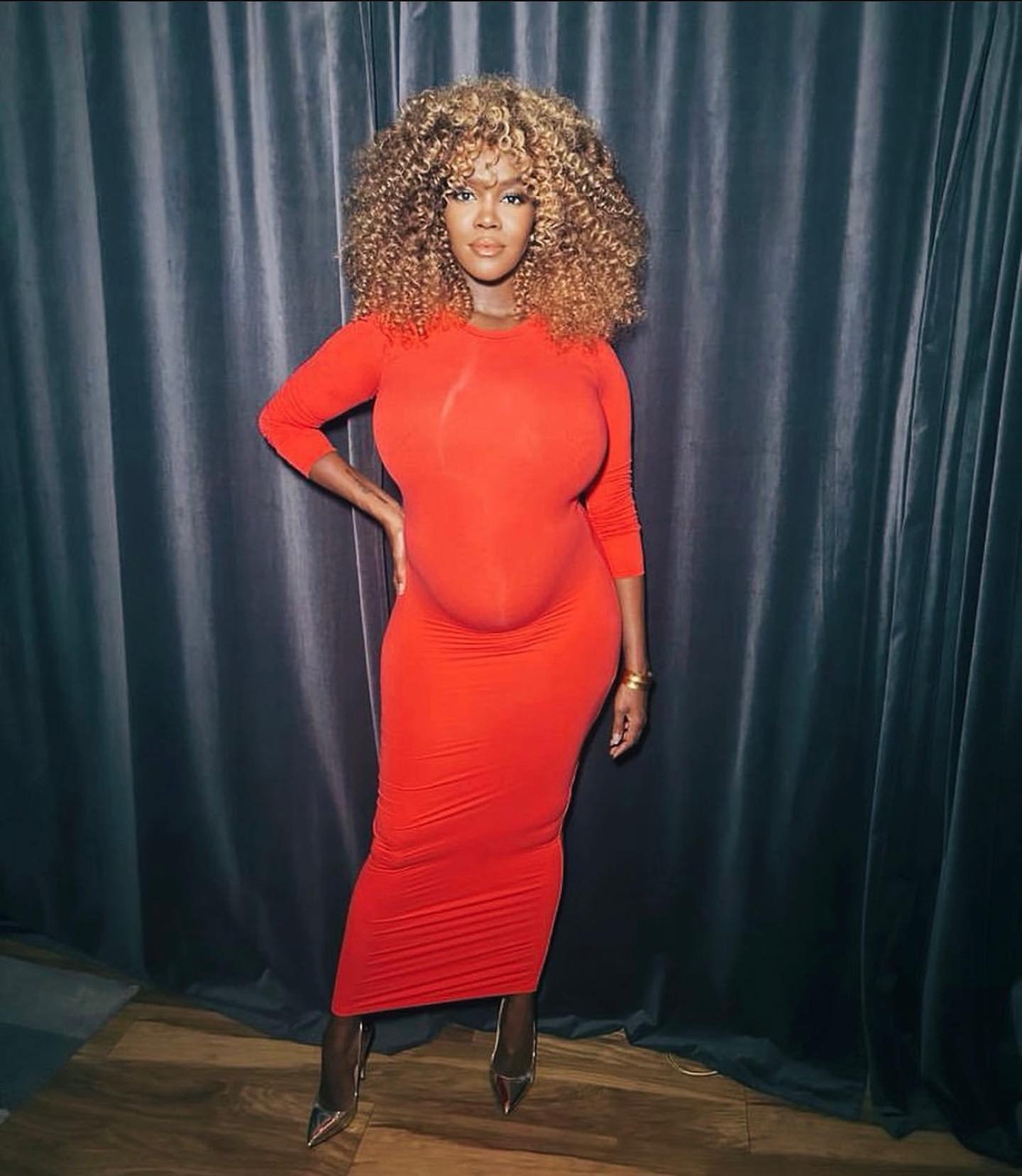 Woman in a long-sleeved orange dress with curly blond hair poses against a grey draped backdrop, showcasing fashion and style.