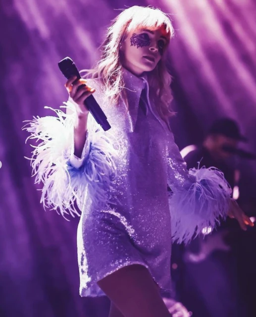 A female singer on stage performs with a microphone, wearing a sequined dress with feather trim and unique eye makeup under purple lighting.