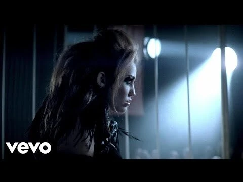 Adult woman with dark hair and dramatic eye makeup looks off-camera in a music-video style shot with blue light and metal bars.