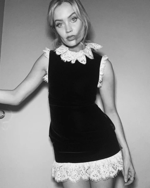 Portrait of a young adult woman in a black dress with white lace collar and hem, against a neutral wall, shot in black and white.