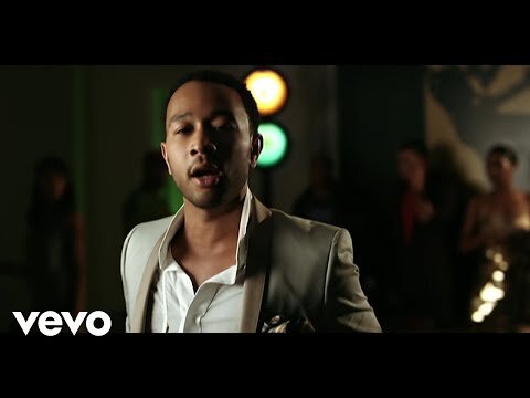 An adult male in a light-colored suit stands in front of blurred figures, with colorful lights in the background, captured in medium shot.