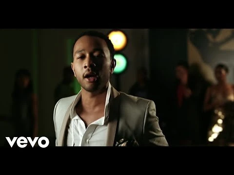 An adult male in a light-colored suit stands in front of blurred figures, with colorful lights in the background, captured in medium shot.