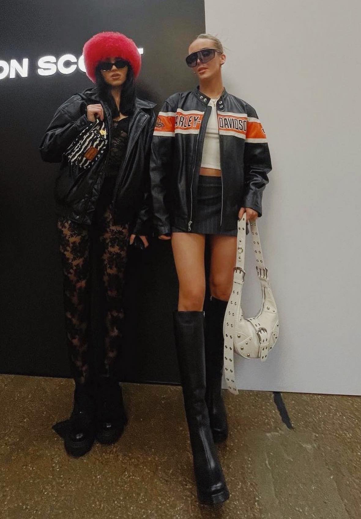 Two women pose in black leather jackets and sunglasses, one with a red furry hat, against a black and white wall. 