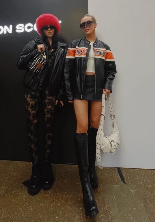 Two women pose in black leather jackets and sunglasses, one with a red furry hat, against a black and white wall. 