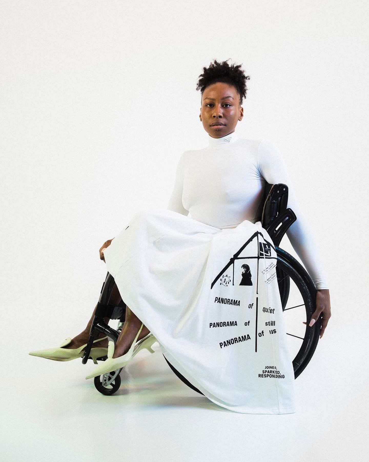 Black woman in white outfit and heels, seated in wheelchair, minimalist backdrop, featuring graphic design on a white drape.