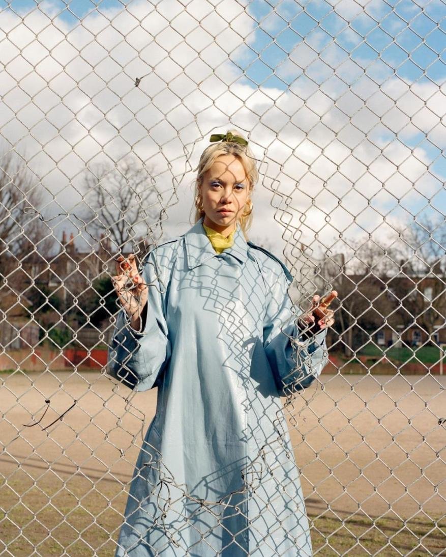 Portrait of a young blonde woman in a light blue coat standing behind a metal chain-link fence against a backdrop of buildings and sky.
