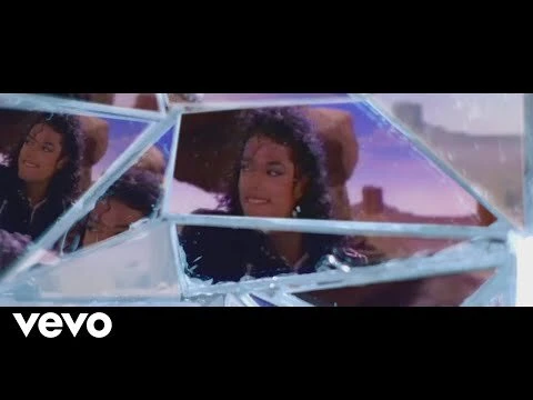 Adult male in black jacket reflecting in broken mirror in desert setting, looks off to the side in a music video scene.