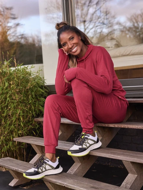 A smiling African-American woman in a maroon sweatsuit sits on wooden steps outside, near greenery, wearing black and white sneakers.