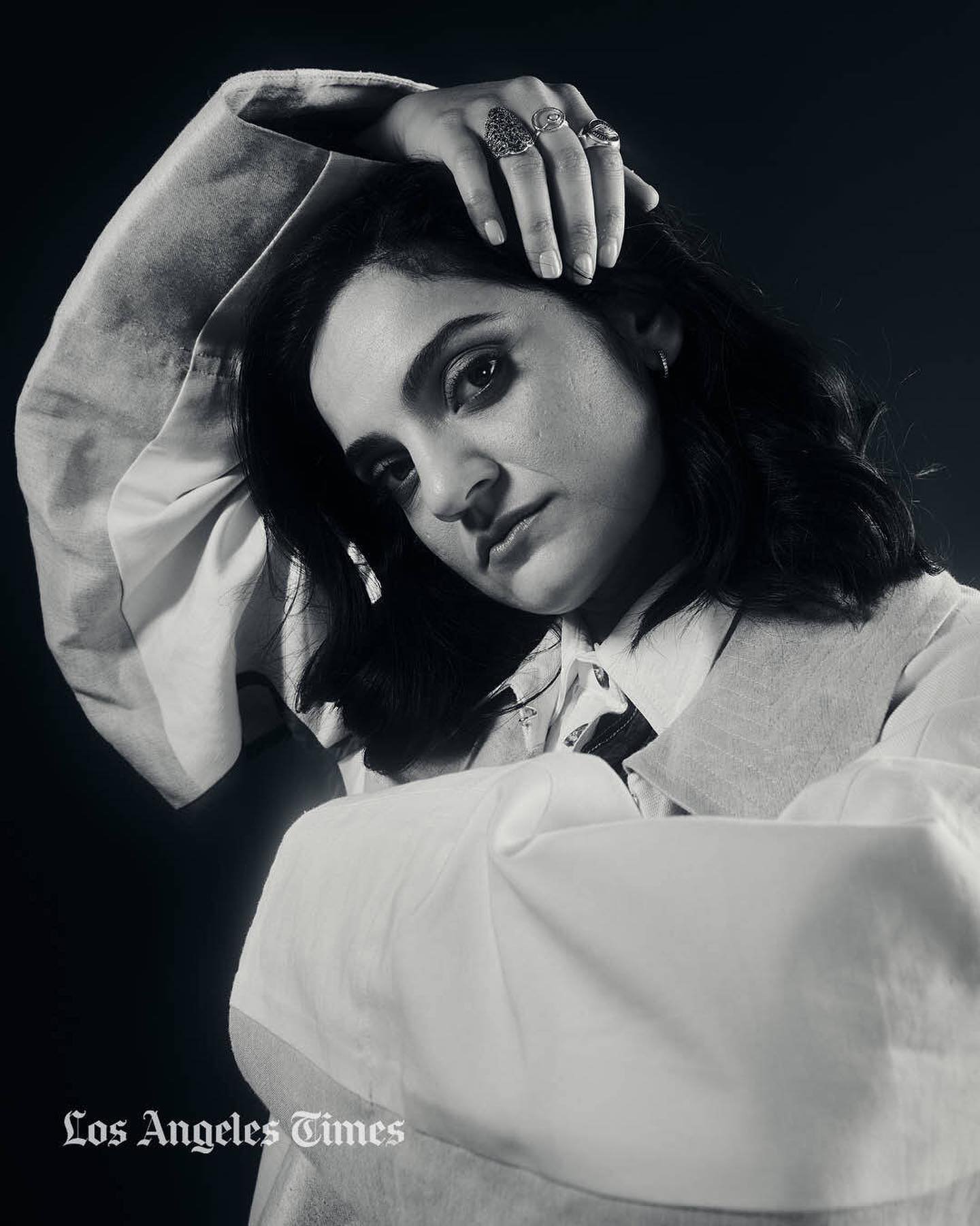 Monochrome portrait of a woman with dark hair wearing rings and a layered white shirt against a dark background.