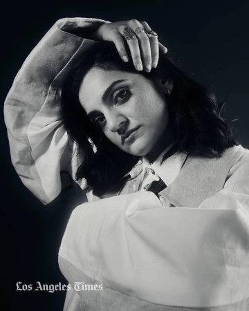 Monochrome portrait of a woman with dark hair wearing rings and a layered white shirt against a dark background.