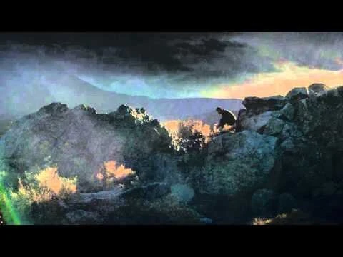 Silhouette of a person crouching on rocky terrain with mountains and a dramatic sky in the background during dusk or dawn.