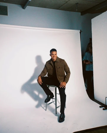 Man seated on a stool in a studio wearing a brown jacket and dark trousers