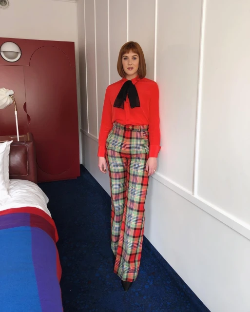 A woman in a red blouse and plaid pants poses indoors with natural light. She has short red hair and a black bow tie.