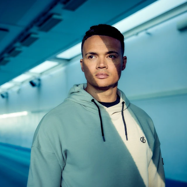 Portrait of an adult male wearing a two-tone hoodie in a blue-toned tunnel with natural light casting shadows across the subject's face.