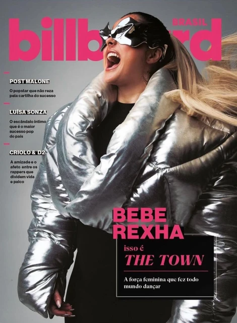 Bebe Rexha on Billboard Brasil magazine cover. She wears a silver puffy jacket and futuristic silver glasses against a pink background.
