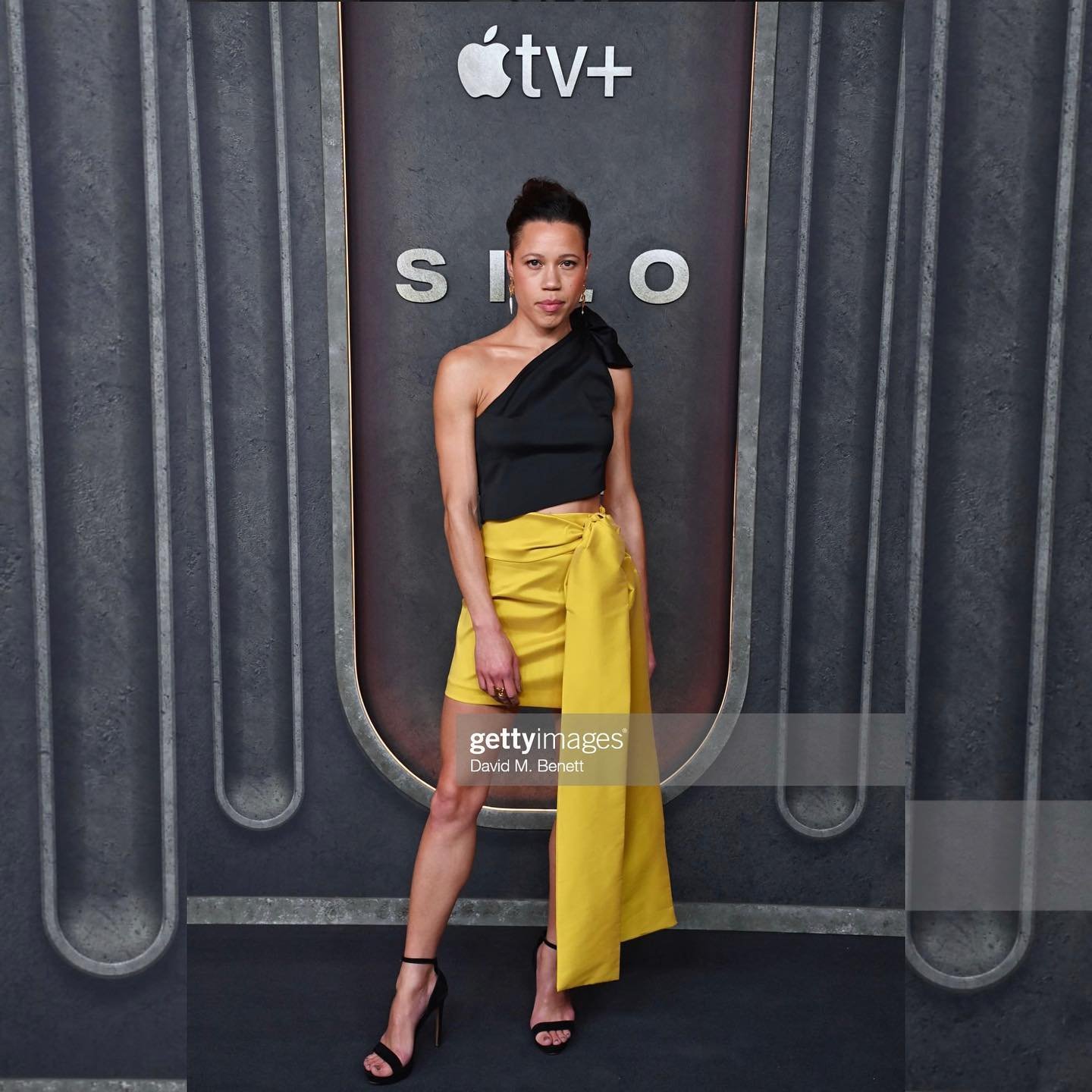 A woman in a black top and yellow skirt poses in front of a 'SILO' backdrop, wearing high heels and accessorized with earrings, at an event.