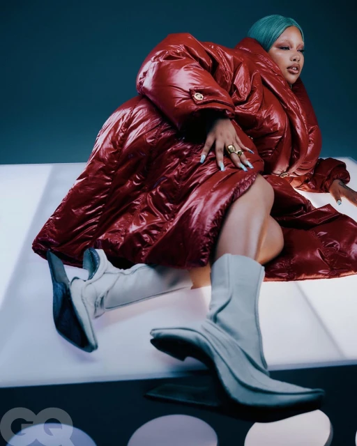 Person in a red puffer coat and white boots laying on a white surface with polka dots and a green-blue background in studio lighting.