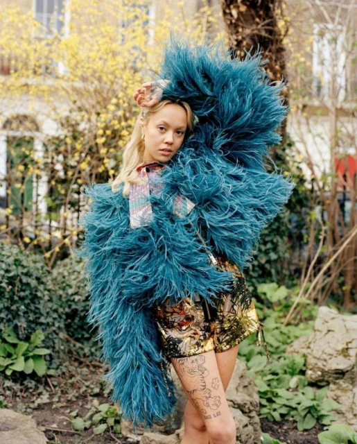 Young woman in statement blue feather coat standing in a garden with trees and yellow flowers on a sunny day.