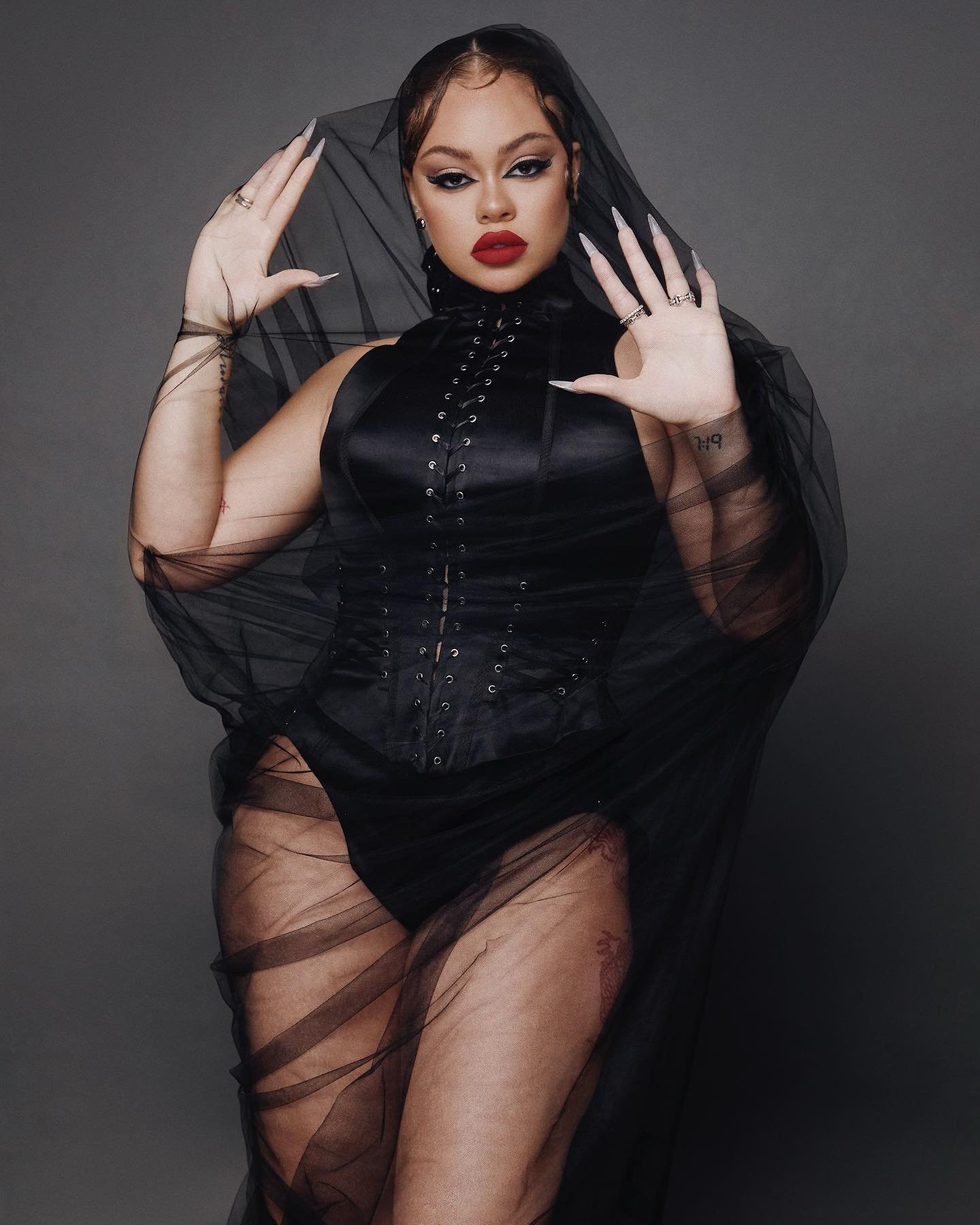 A woman poses in a black bodice and veil, adorned with rings and nail extensions, against a grey backdrop in a fashion portrait.