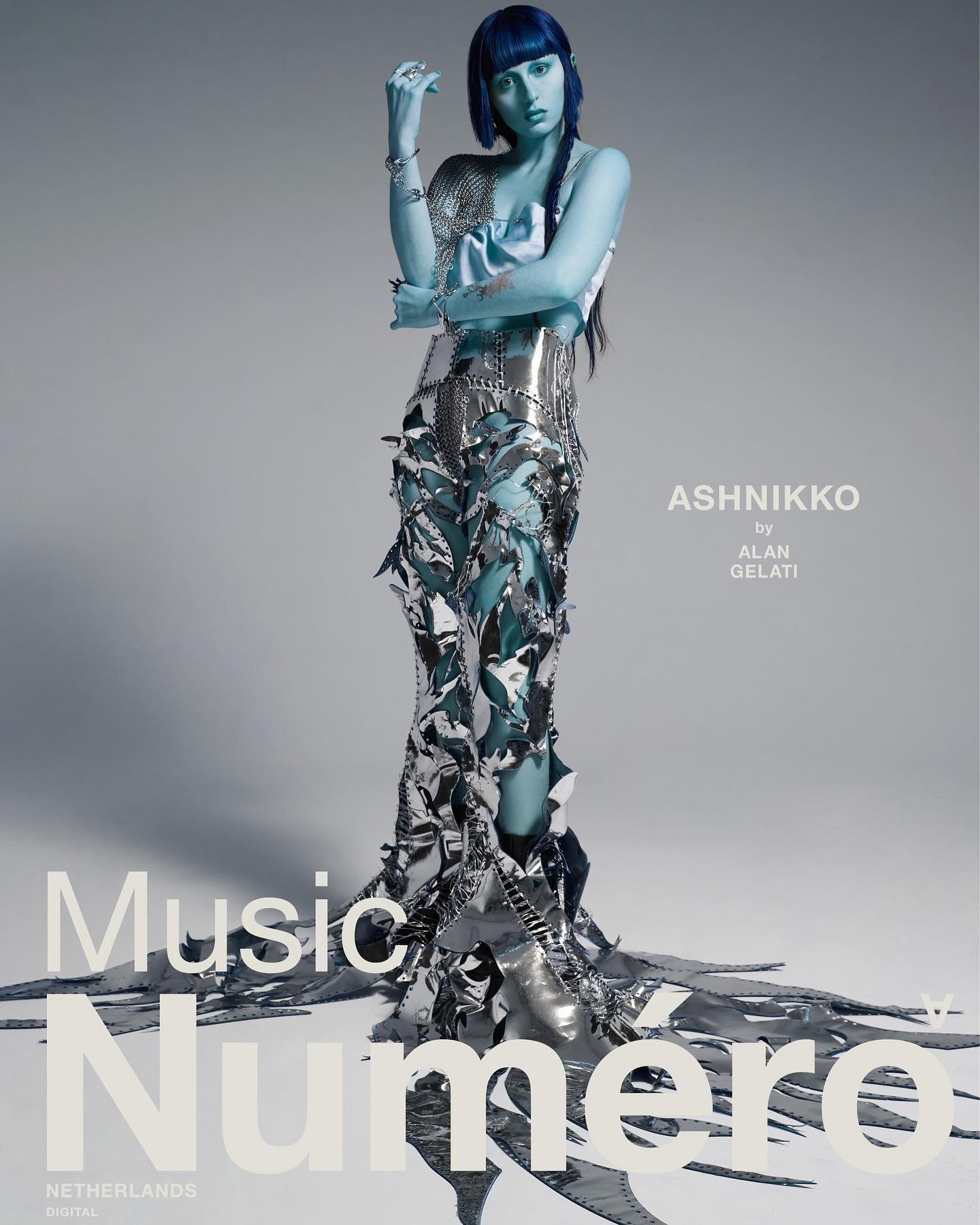 Model with blue-painted skin and dark hair posing in a sculptural metallic outfit against a grey background