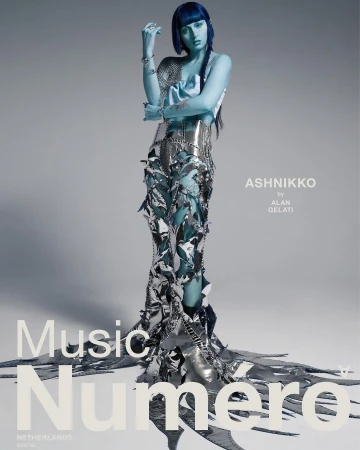 Model with blue-painted skin and dark hair posing in a sculptural metallic outfit against a grey background
