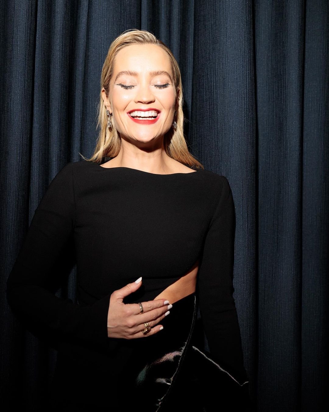 Blonde woman in a black long-sleeved dress smiles with her eyes closed in front of a textured dark blue curtain backdrop.