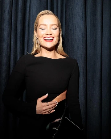 Blonde woman in a black long-sleeved dress smiles with her eyes closed in front of a textured dark blue curtain backdrop.