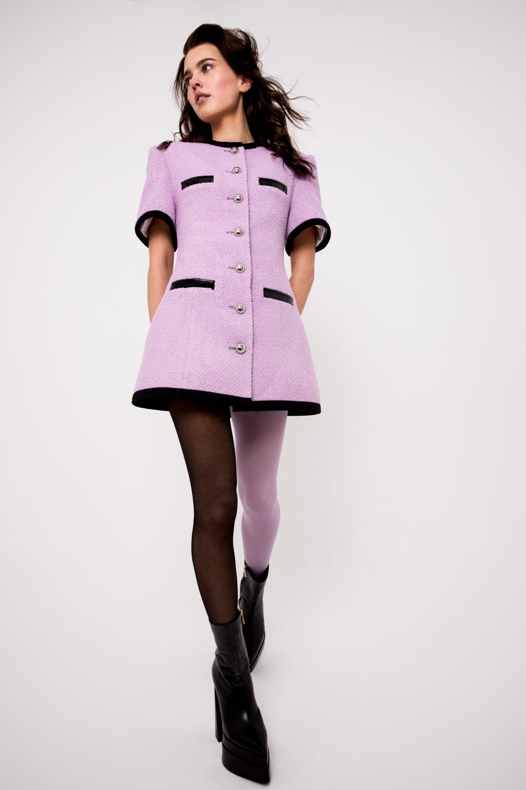 Elegant woman in a lilac tweed dress with black trim and black ankle boots, posing against a white backdrop in a fashion shot.