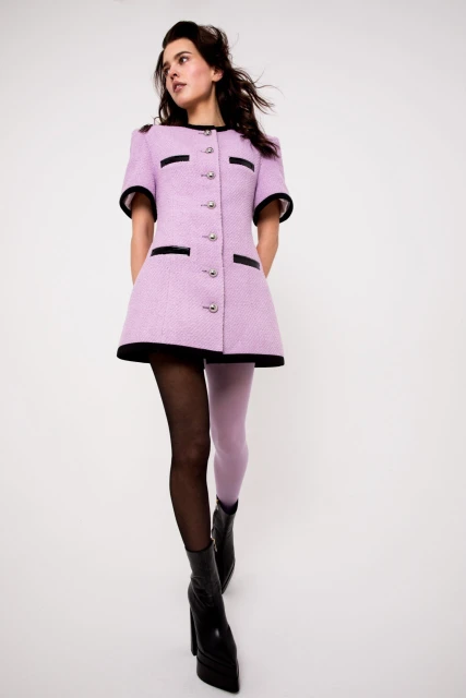 Elegant woman in a lilac tweed dress with black trim and black ankle boots, posing against a white backdrop in a fashion shot.
