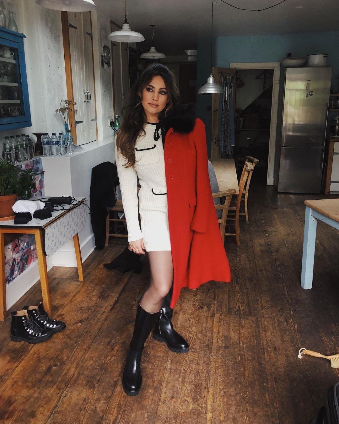 A woman with long brown hair poses indoors wearing a white dress and knee-high boots, holding a red coat, in an interior space.